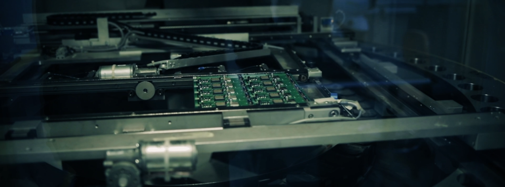 A close-up of a mechanical assembly with a green printed circuit board on a conveyor system, surrounded by metal components.
