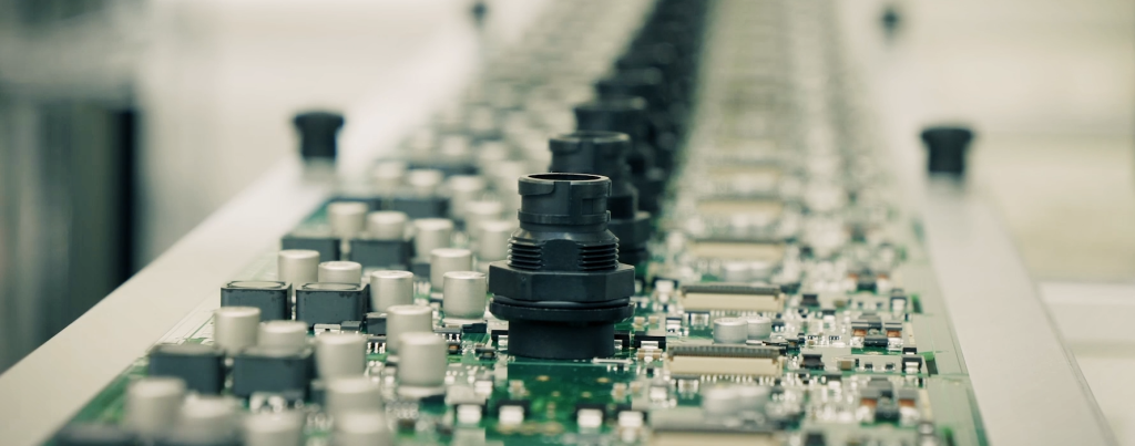 A close-up of circuit boards with various components, including metallic caps and connectors, arranged in a production line setting.